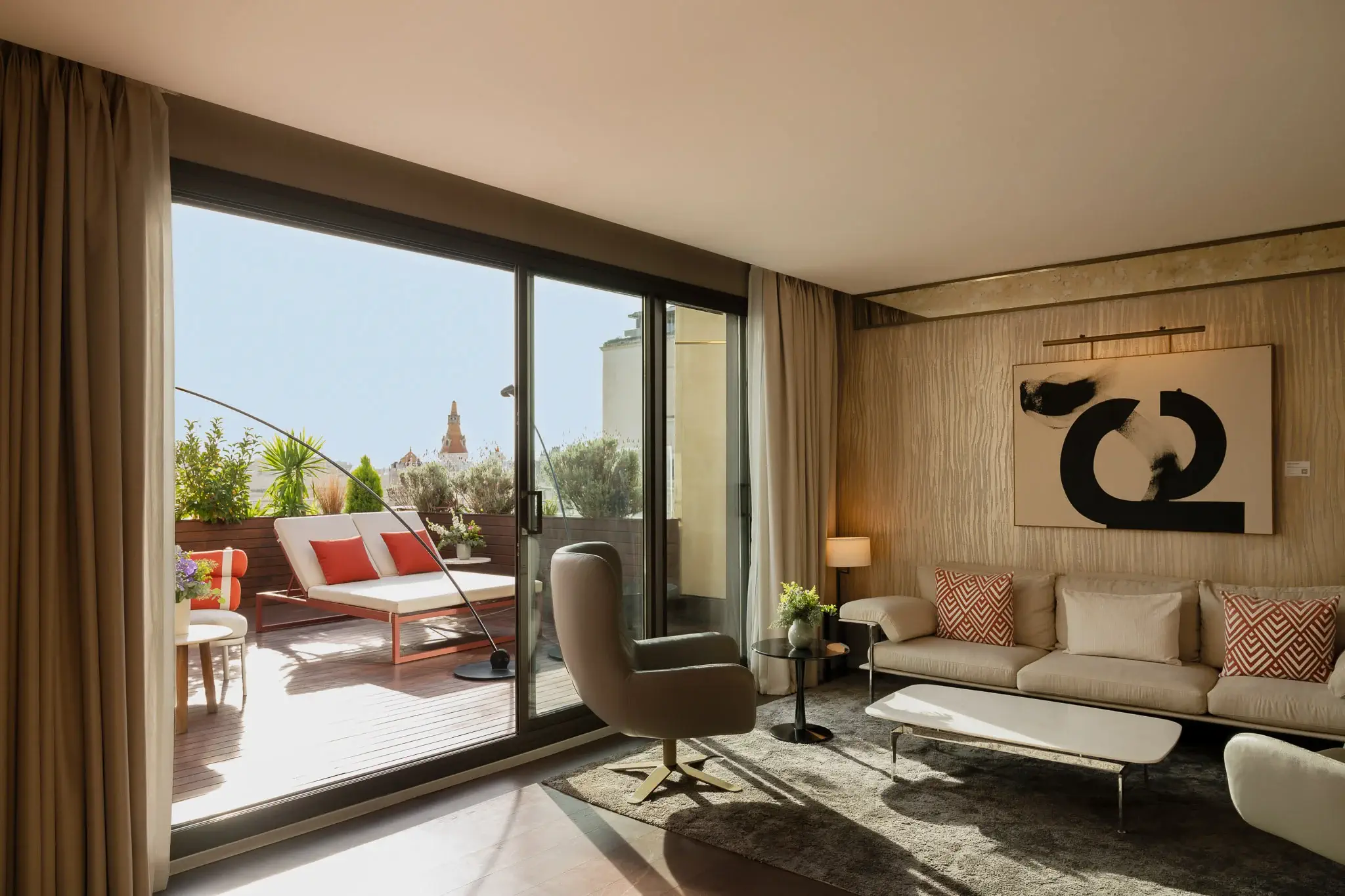 Una moderna sala d'estar tipus suite Grand Penthouse amb tons neutres, art mural abstracte i portes de vidre que s'obren a un pati assolellat amb seients a l'aire lliure i plantes exuberants.