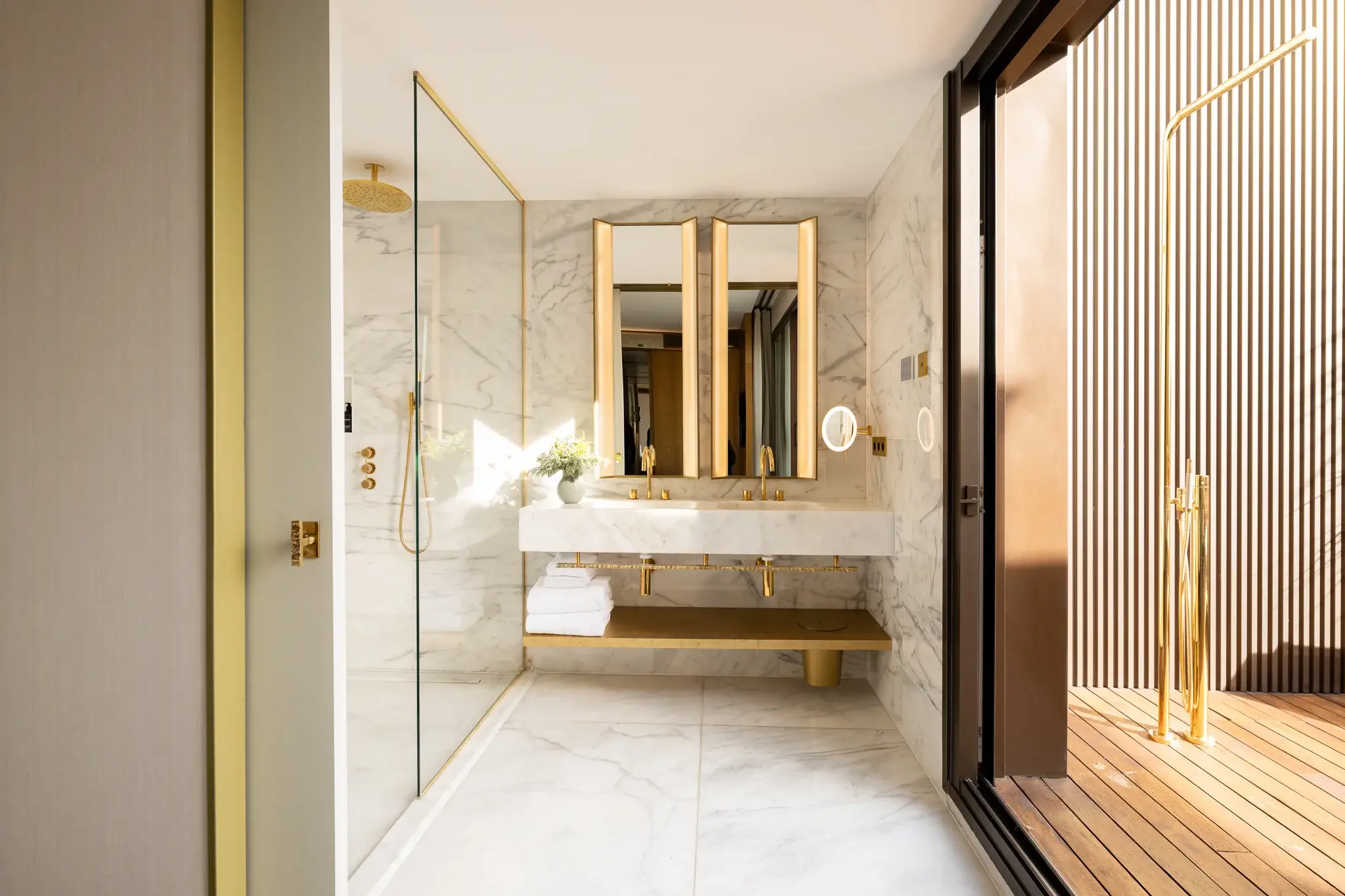 Bany modern a la suite Grand Penthouse amb parets i terres de marbre, accessoris daurats, lavabo doble, miralls grans, dutxa de vidre i una porta corredissa que s'obre a una zona de terrassa de fusta.