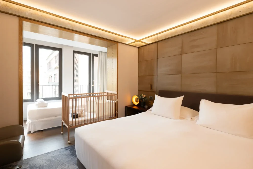 A modern hotel room with a large double bed, a wooden crib, a single bed by the window, and floor-to-ceiling windows providing natural light.