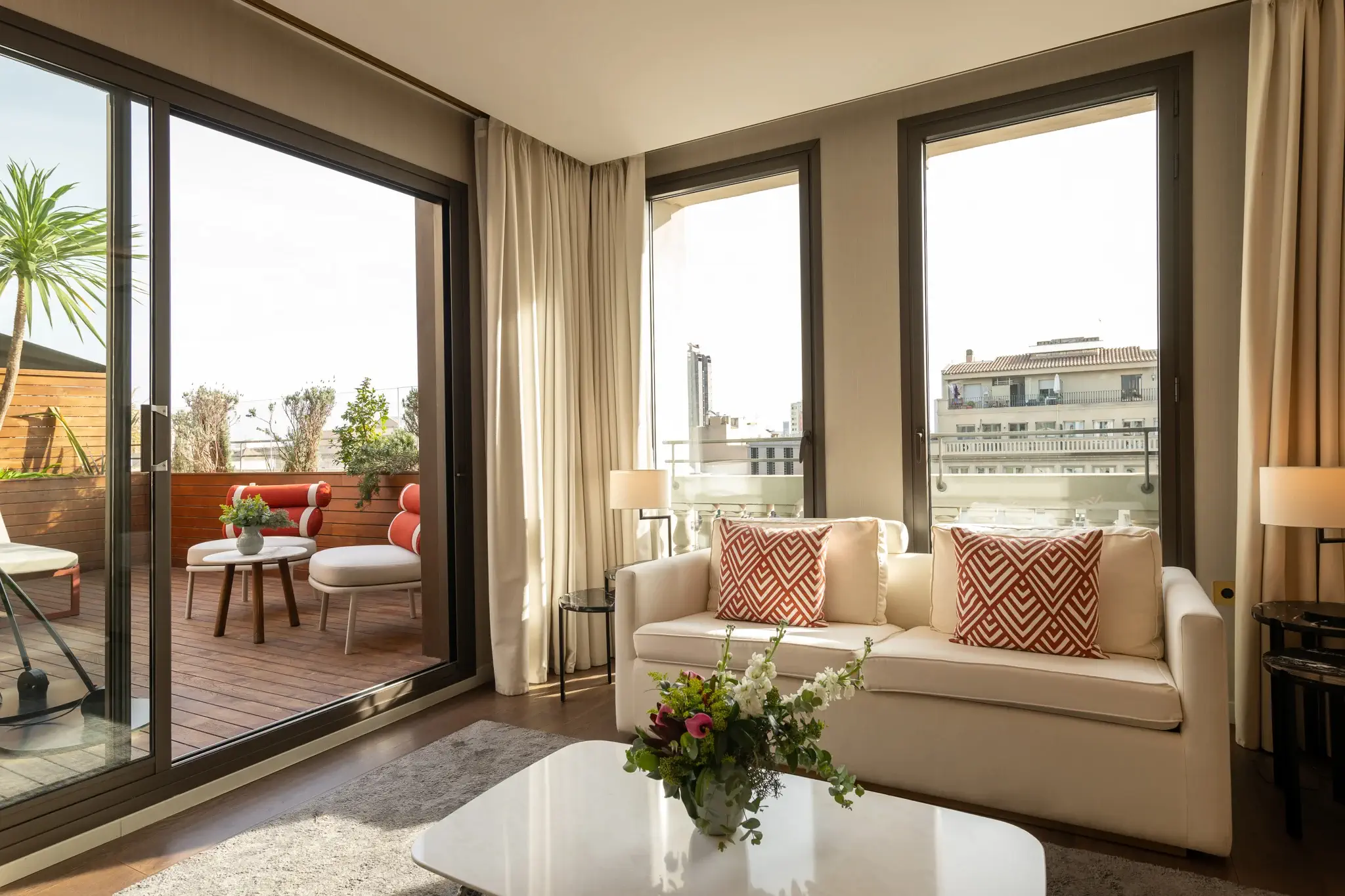 Modern living room with a white sofa, patterned red cushions, large windows, and a view of the furnished Terrace Suite. Natural light fills the space.