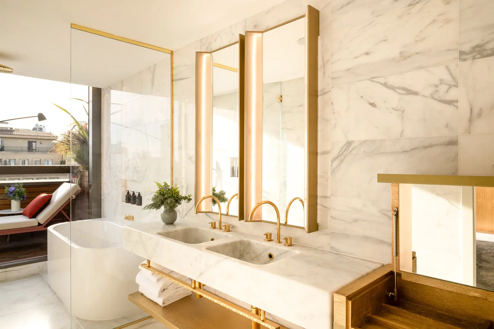 Modern Terrace Suite bathroom with marble walls, double sink vanity with gold fixtures, large mirrors, a freestanding bathtub, and an outdoor seating area visible through glass doors.