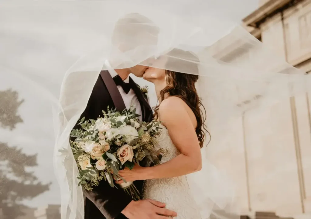 A bride and groom kiss under a sheer veil outdoors at Almanac Barcelona, with the bride holding a bouquet of flowers and the groom wearing a black suit.