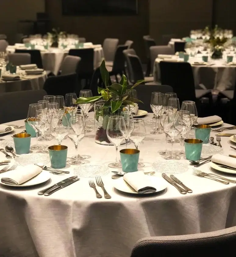 A round table set for formal events or meetings features multiple glasses, silverware, teal cups with gold rims, white napkins, and a small green centerpiece. Other similarly set tables are in the background.