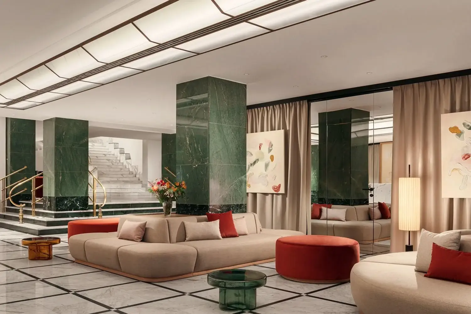 Moderní hotelová hala hotelu Almanac Alcron Prague s béžovými pohovkami, červenými otomany, zelenými mramorovými sloupy, abstraktním uměním a schodištěm s mosazným zábradlím v pozadí.