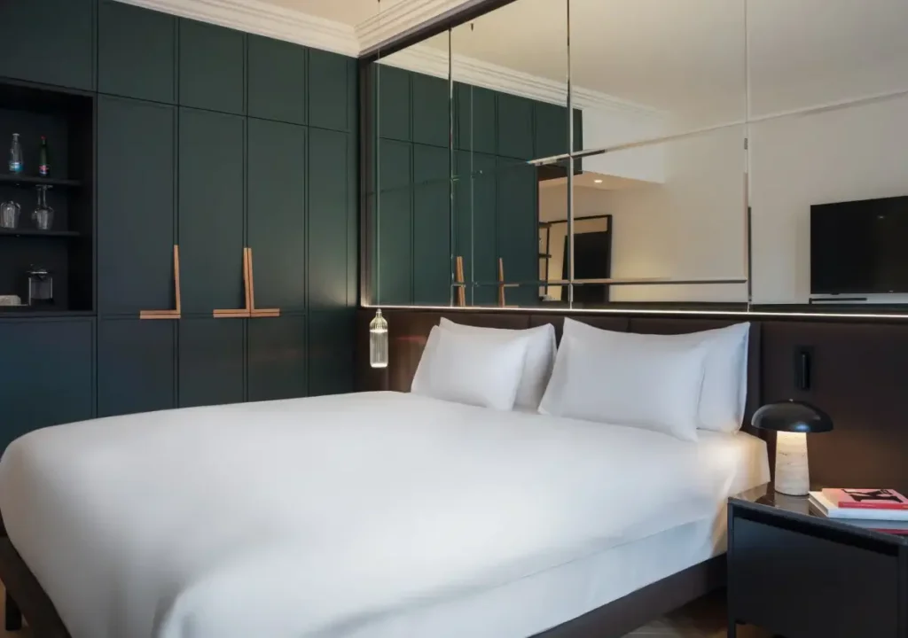 A modern hotel room at Alcron Prague features a neatly made double bed, a dark bedside table with a lamp, wall-mounted shelves, and large mirrors above the headboard.