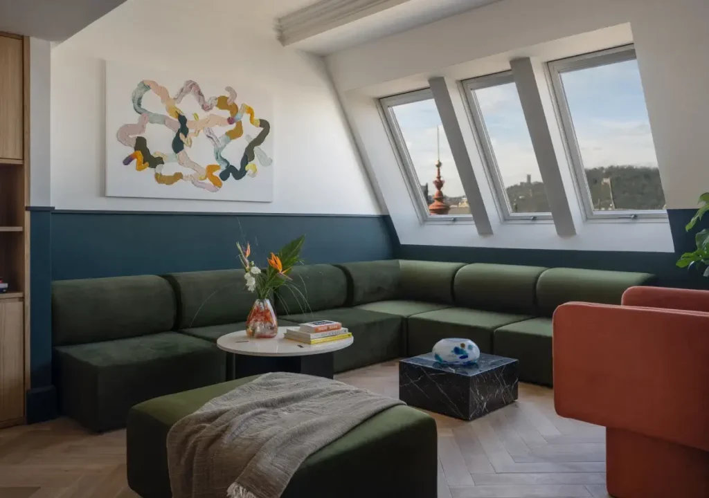 Modern living room with green sectional sofa, abstract wall art, angled windows, and a round coffee table adorned with flowers and books—an inviting space reminiscent of the stylish lounges at Almanac or Alcron Prague.