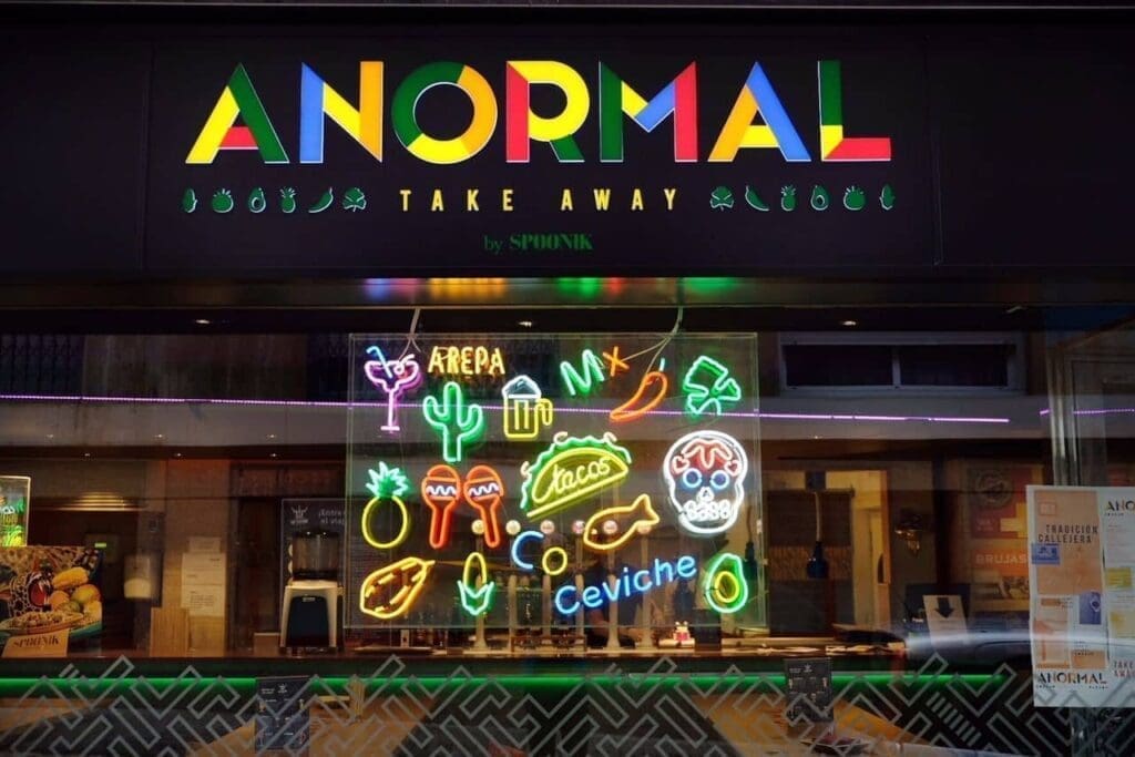 Storefront with an "ANORMAL TAKE AWAY" sign and neon window display featuring tacos, cactus, skulls, chili peppers, and words like “Arepa” and “Ceviche”—a vibrant showcase of Anormal Food.