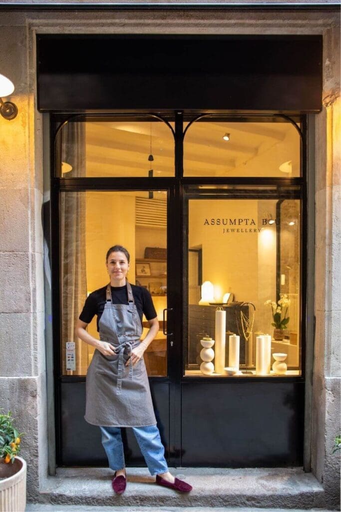 A person wearing an apron stands in front of a jewelry shop with glass windows displaying decorative items and the sign "ASSUMPTA B JEWELLERY" by Assumpta Bou visible behind them.