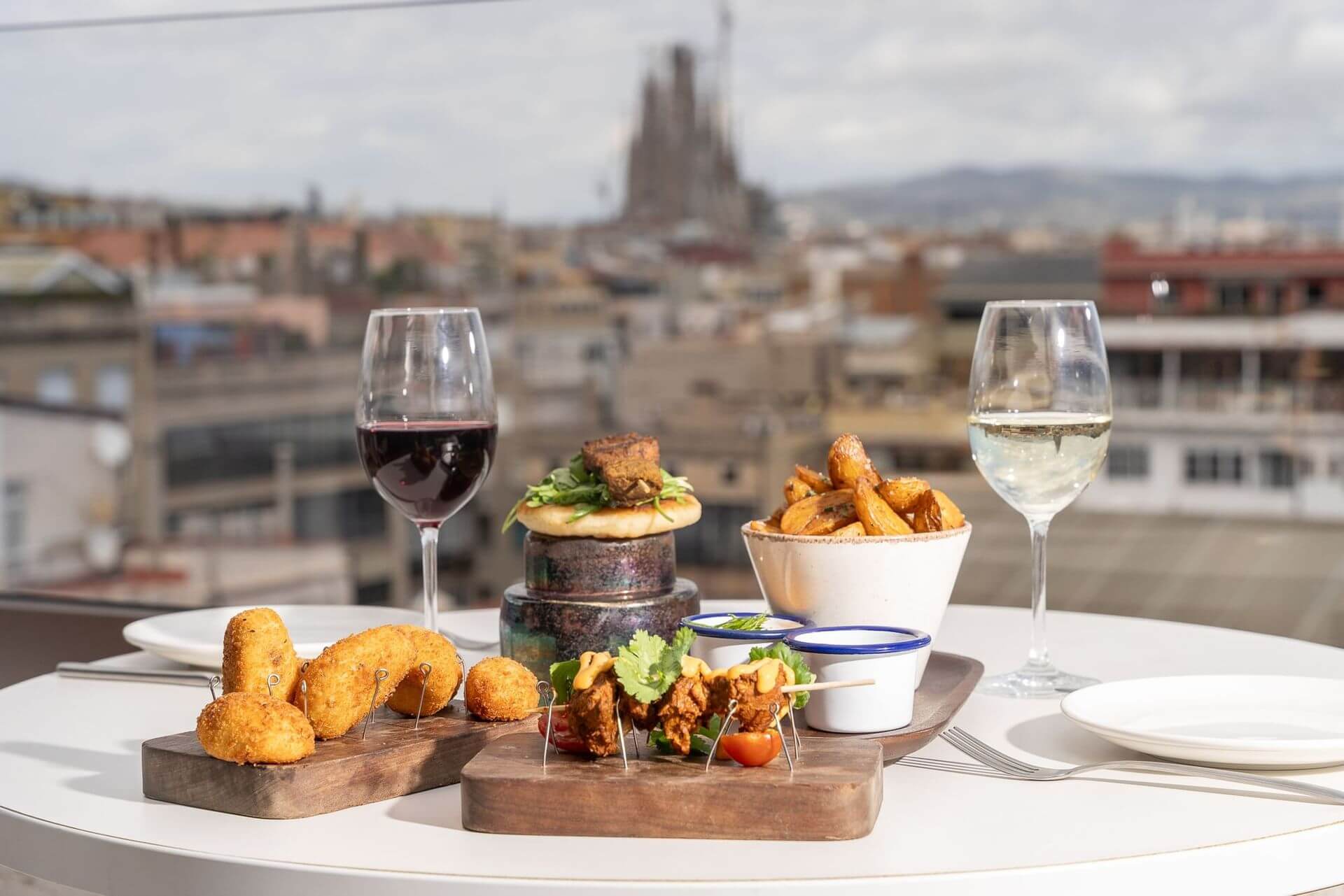 Una taula parada amb tapes variades, un bol de patates fregides i copes de vi negre i blanc en un terrat de Terrassa, amb vistes al paisatge urbà i la catedral sota el cel clar d'Azimut.