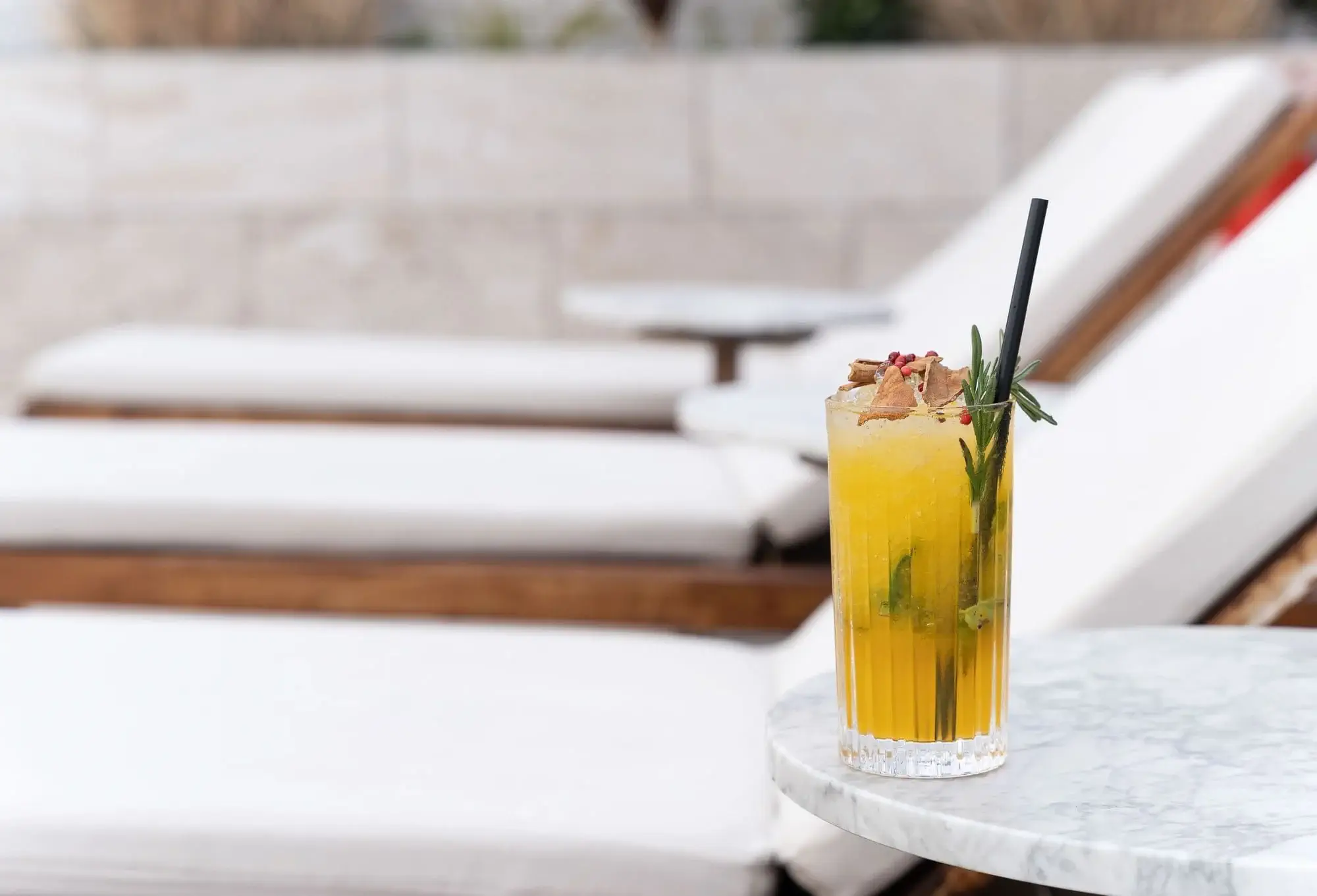 Un còctel groc amb gel, herbes i una palla negra es troba sobre una taula rodona de marbre al costat de butaques blanques a la terrassa, capturant perfectament l'ambient relaxant de la piscina.