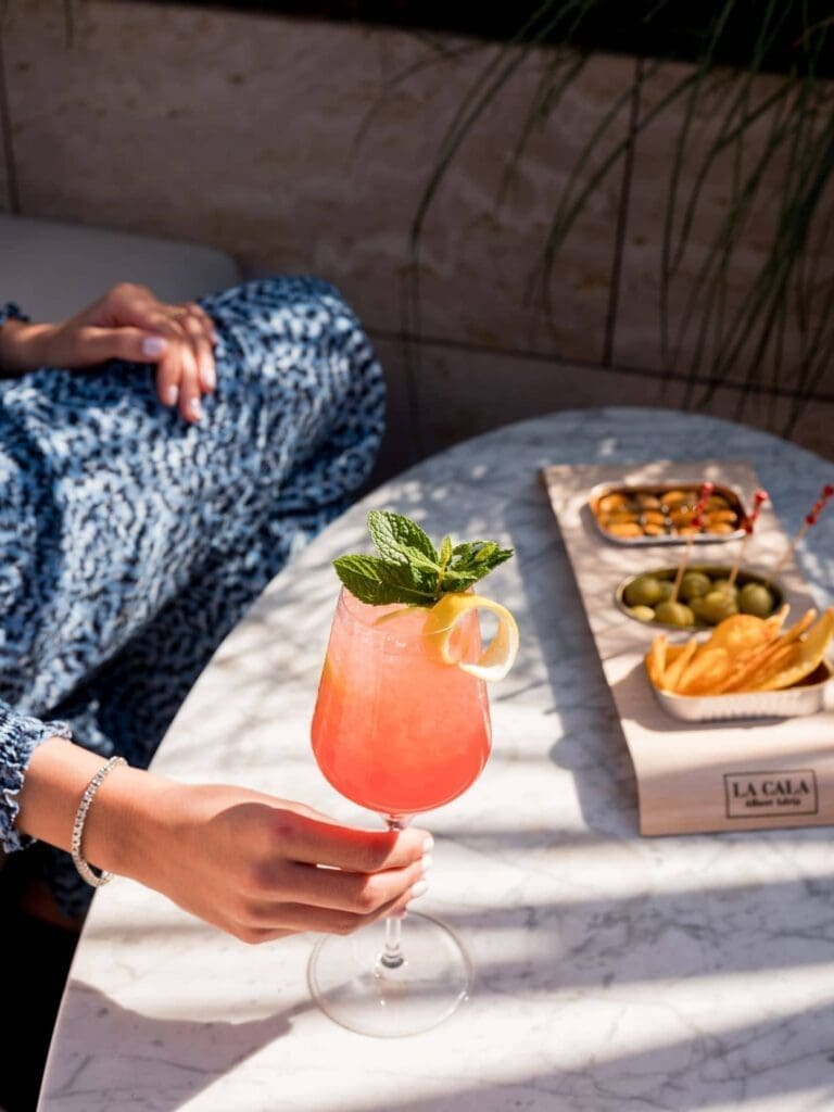 A person holds a pink cocktail garnished with mint and lemon at a marble table at Azimuth Rooftop Bar, surrounded by bowls of olives, chips, and nuts in sunlight.