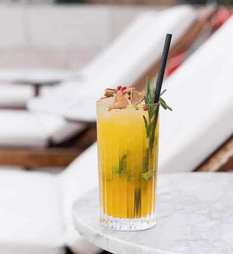 A yellow cocktail with ice, herbs, and garnish is served in a tall glass with a black straw on a marble table beside white lounge chairs at Azimuth Rooftop Bar.