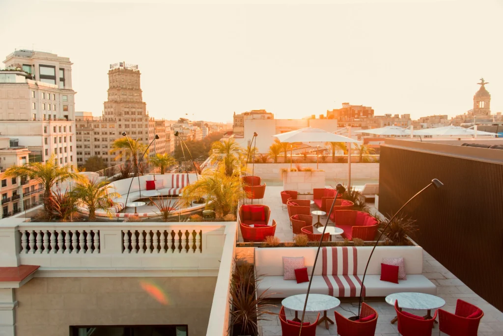 Die Dachterrasse des Almanac Barcelona mit ihren rot-weißen Sitzgelegenheiten, Topfpflanzen und Sonnenschirmen bietet einen Blick auf das Stadtbild bei Sonnenuntergang.