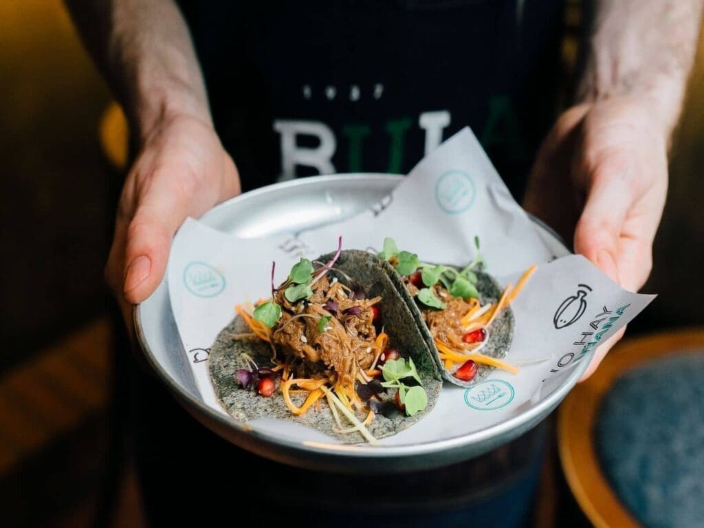 A person holds a plate with two tacos filled with shredded meat, microgreens, shredded vegetables, and pomegranate seeds, served on Babula Bar 1937 branded paper.