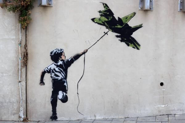 L'art urbà de BANKSY captura l'art de la protesta, representant un nen que sosté una corda lligada a un globus amb forma de jet militar sobre un mur de formigó.