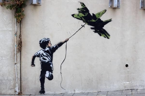 Street art inspired by Banksy: a child holds a string tied to a military fighter jet, floating like a balloon, symbolizing Art of Protest on a beige wall.