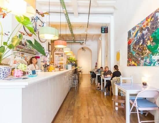 Bright, modern café with a long white counter, colorful hanging lights, and several people seated at tables along the wooden floor.