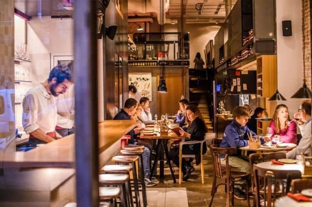 People dining and socializing in a modern restaurant with a chef preparing food behind the counter on the left.
