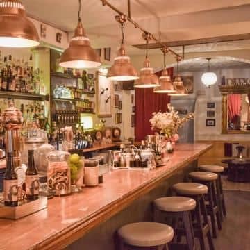 A cozy bar with a wooden counter, high stools, hanging pendant lights, and shelves stocked with bottles and glassware serving signature cocktails made with Dux Gins.