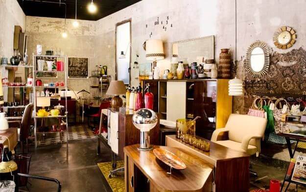 A vintage shop interior with mid-century furniture, shelves of decorative items, lamps, clocks, and handbags displayed against a worn, textured wall reminiscent of Barcelona’s Montjuïc Castle or a historic fortress.