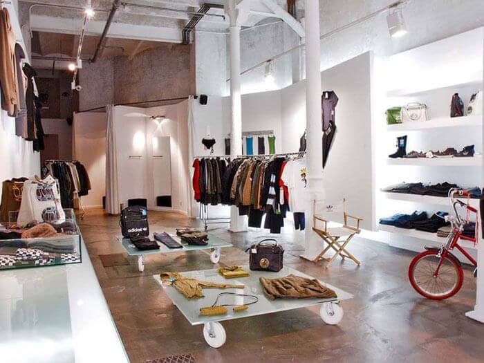 Clothing store interior in Barcelona with racks of clothes, shelves of bags and accessories, a red bicycle, and a table displaying folded garments on a polished concrete floor—just steps from the historic Montjuïc Castle.