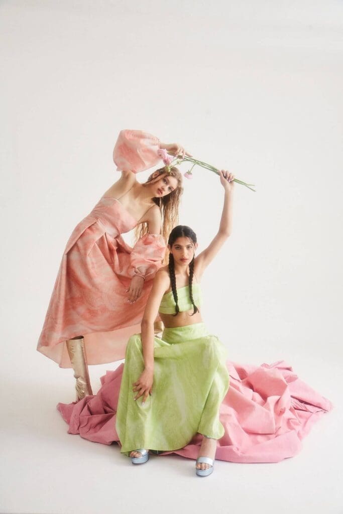 Two women in pastel dresses pose against a white background, evoking the elegance of Barcelona. One stands in a pink dress holding a flower, while the other sits in green with pink fabric draped around her, reminiscent of scenes near Montjuïc Castle.