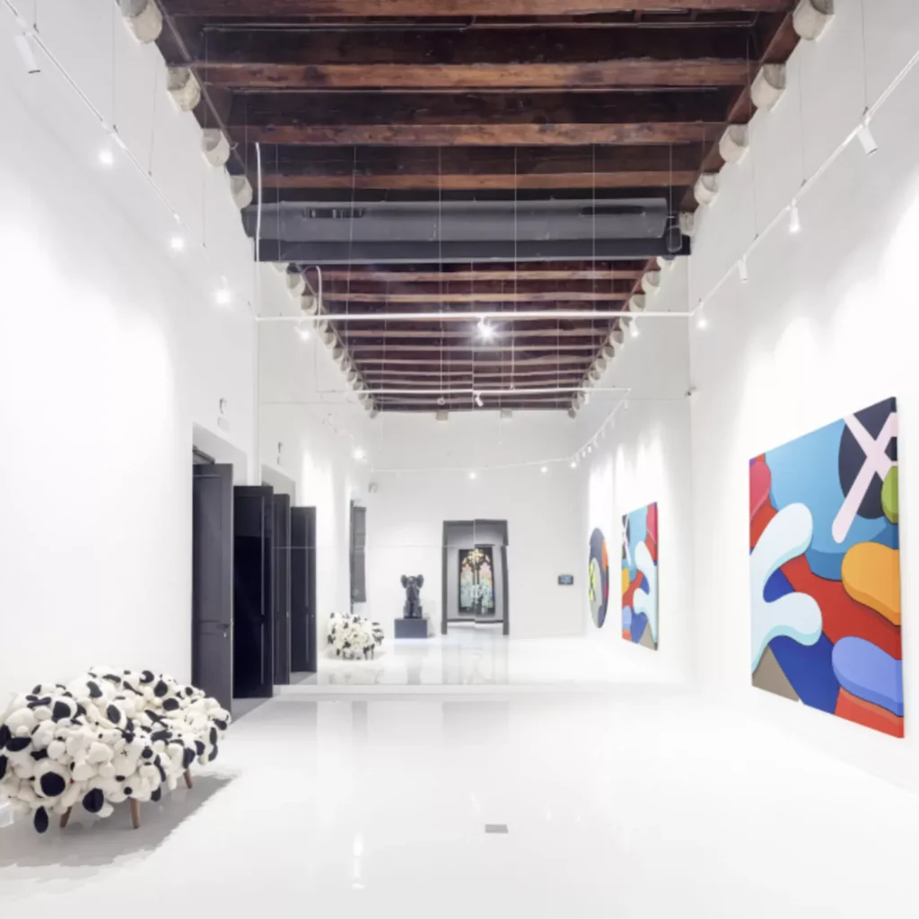 Bright, modern art gallery with white floors and walls, colorful abstract paintings, a black-and-white patterned bench, exposed wooden ceiling beams—perfect for discovering contemporary art inspired by the vibrant spirit of Barcelona.