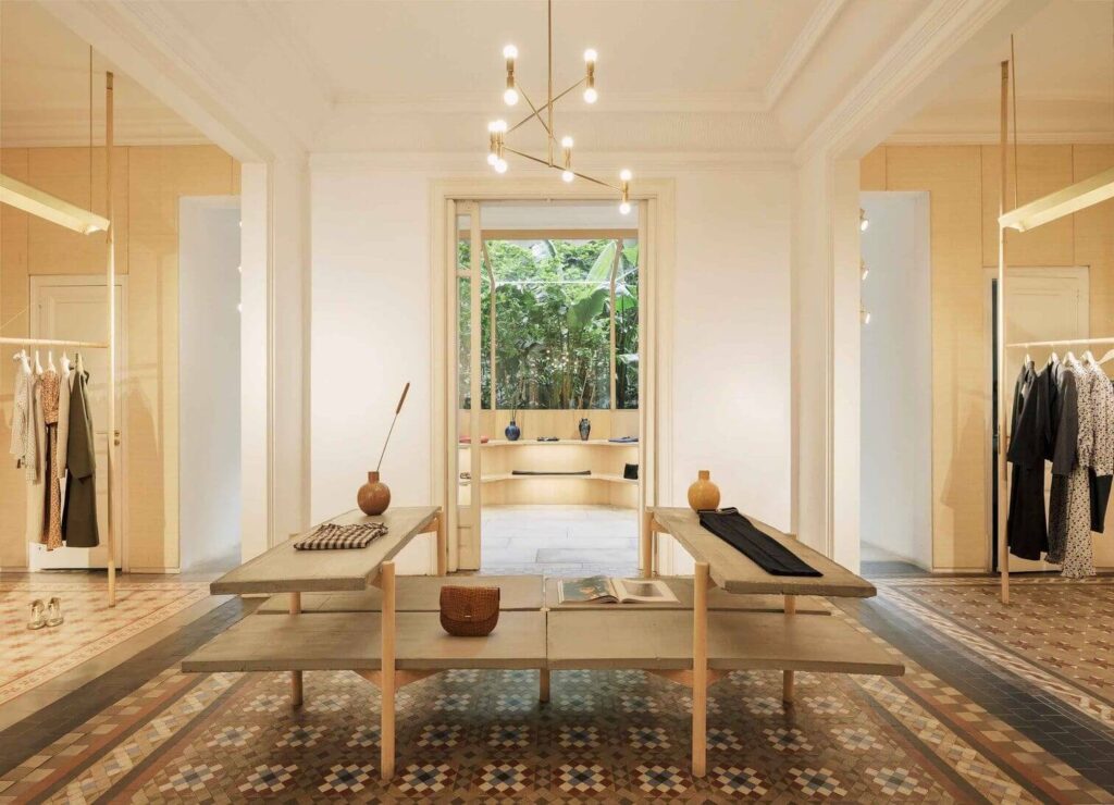Interior de boutique minimalista con suelo de baldosas estampadas, dos mesas de exposición, lámparas colgantes, percheros a ambos lados y un gran ventanal que deja entrever una vegetación que recuerda a los jardines del castillo de Montjuïc de Barcelona.