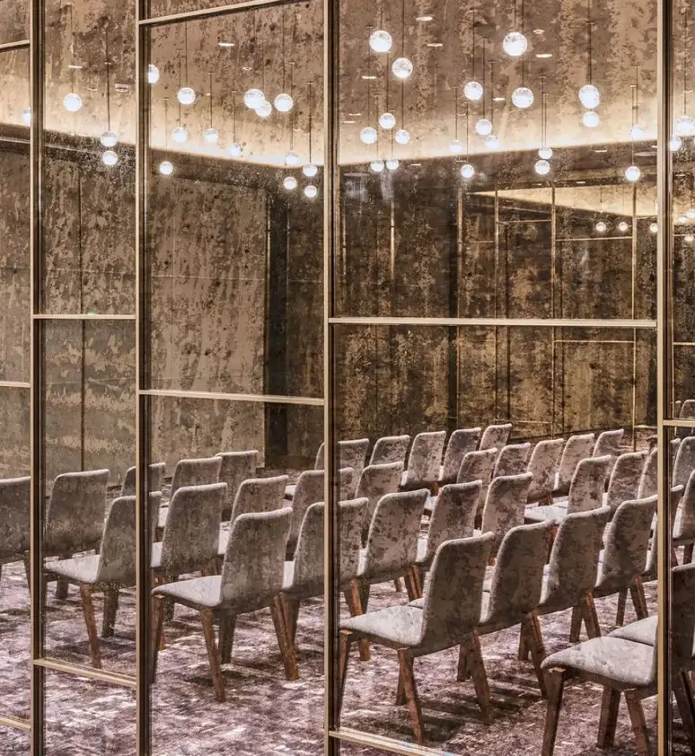 Fileres de cadires buides estan disposades mirant cap endavant en una sala enmoquetada, ideal per a reunions o actes privats, amb parets de miralls i un sostre adornat amb llums rodons penjants.