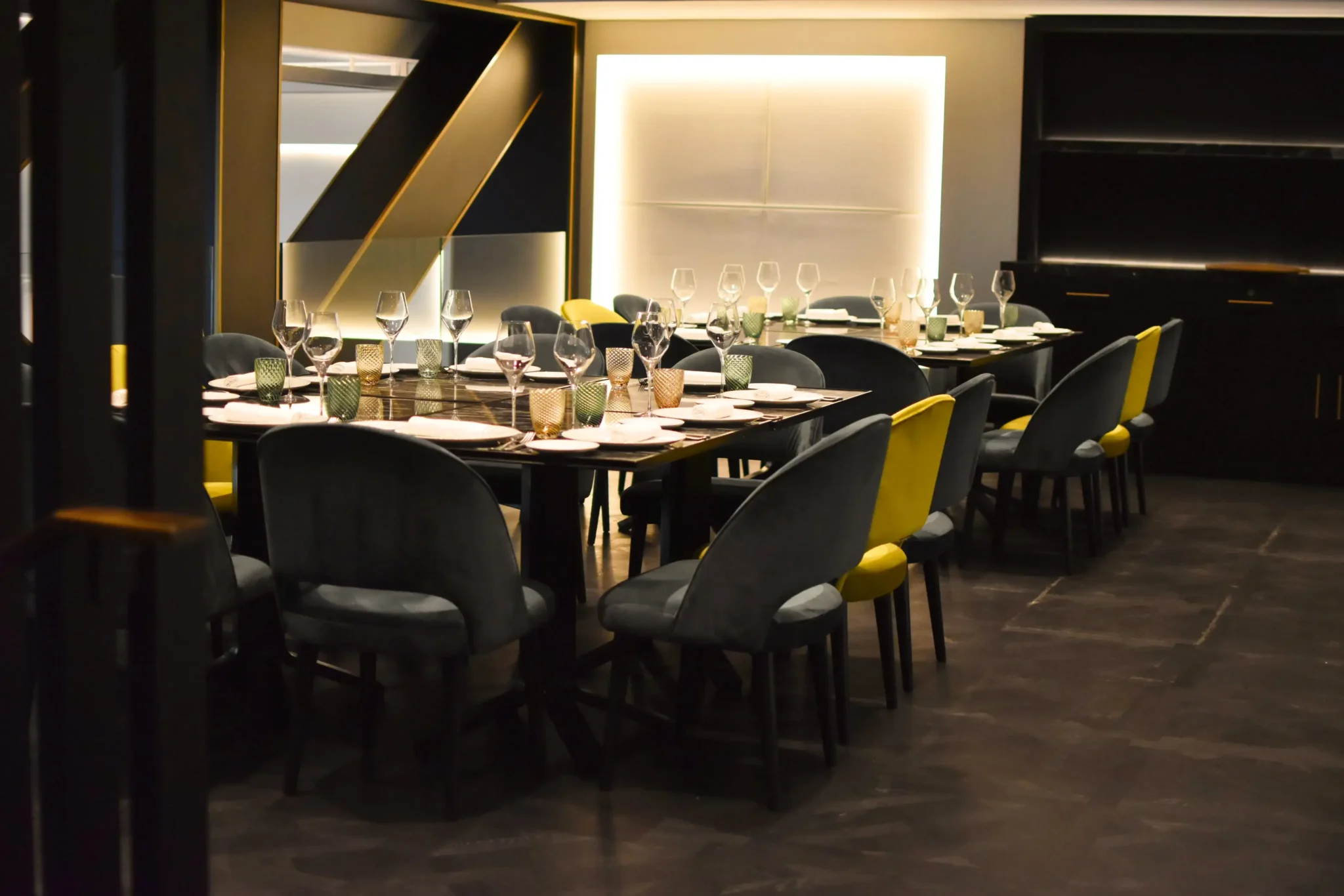 A modern dining room designed for social gatherings, with a long table set for a meal, featuring neatly arranged wine glasses, plates, and cutlery, surrounded by dark and yellow chairs.