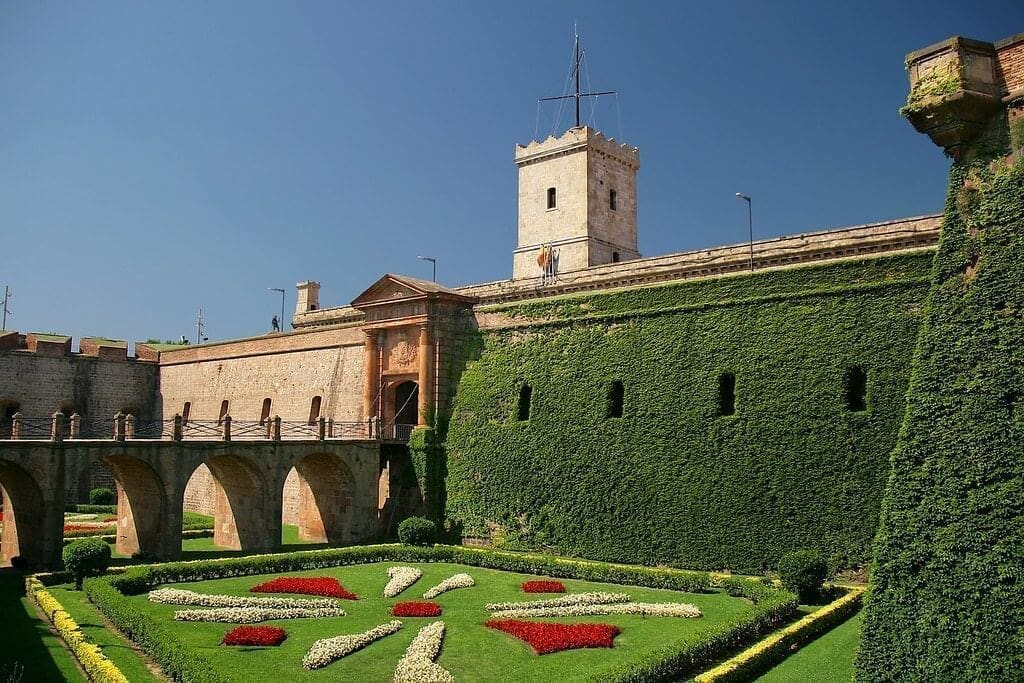 Montjuïc Castle, a historic fortress in Barcelona, boasts ivy-covered stone walls, an arched bridge, and a manicured garden with red and white flowerbeds beneath a clear blue sky.