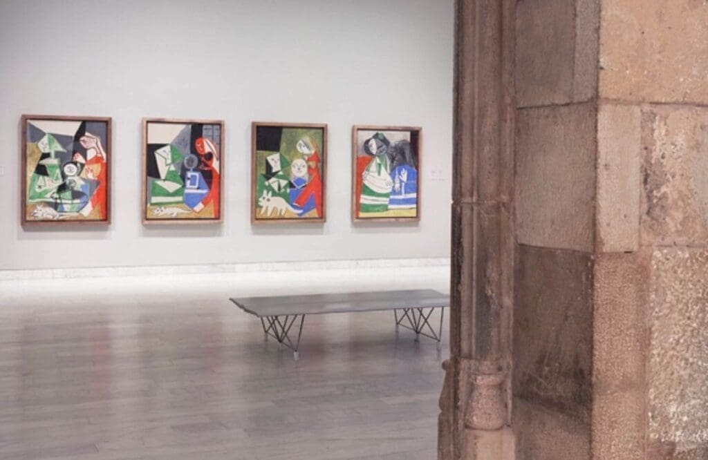 Four abstract paintings hang on a white wall in a gallery at the Picasso Museum, with a bench in front; stone columns are visible in the foreground, echoing the historic charm of Barcelona.