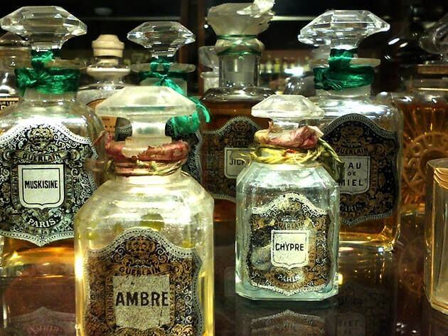 Diversos flascons de perfum vintage ornamentats, etiquetats amb noms com "Ambre", "Chypre" i "Muskisine", es mostren junts sobre una superfície reflectant, evocant l'elegància del famós Museu del Perfum de Barcelona.