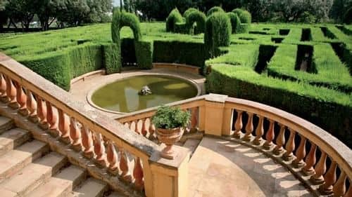 Stone staircase with balustrade leading to a circular pond, surrounded by manicured green hedges forming a labyrinth in the formal Parque Laberinto de Horta garden in Barcelona.
