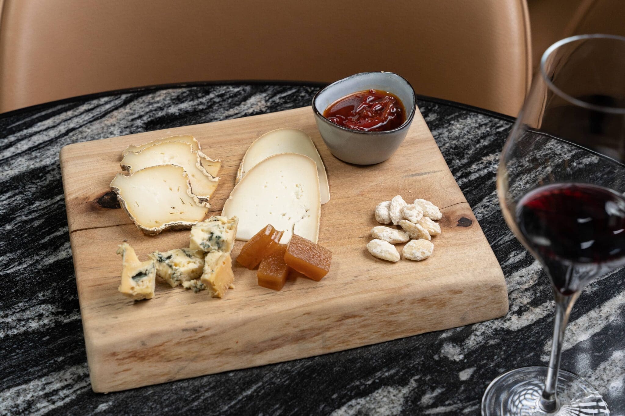 A cheese board with assorted sliced and cubed cheeses, quince paste, almonds, a small bowl of red jam, and a glass of red wine on a dark marble table.