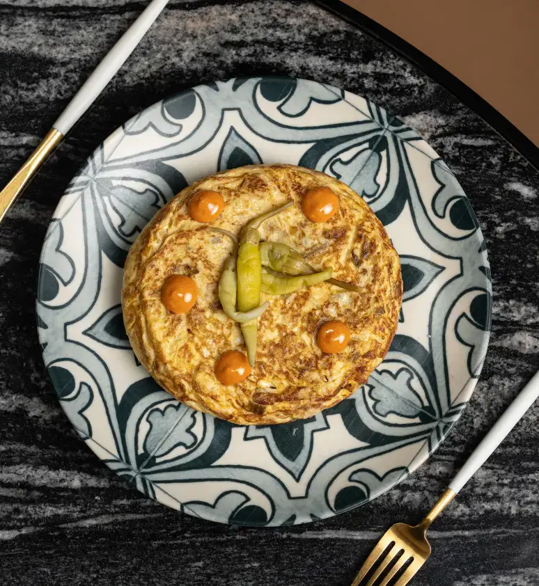 Una truita espanyola amb pebrots verds tallats a rodanxes i cinc cullerades de salsa, servida en un plat estampat amb una forquilla i un ganivet daurats.