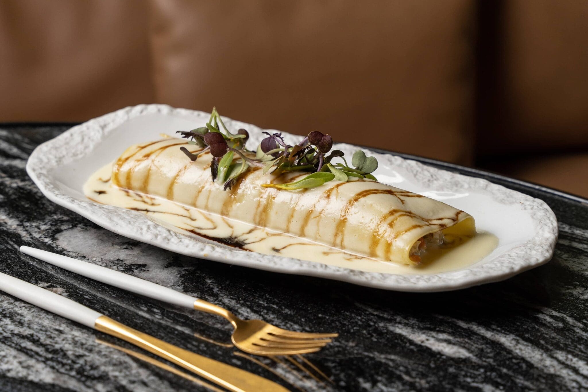 A rolled crepe topped with microgreens and sauce is served on a white plate, accompanied by a fork and knife on a marble table.