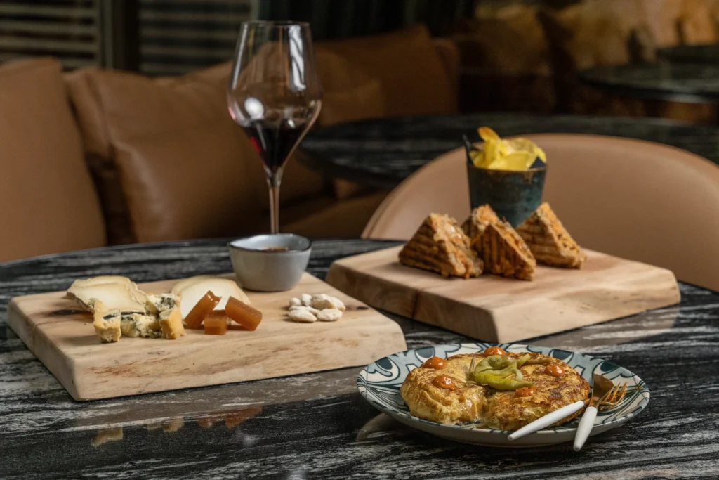 Auf einem Tisch, der für Essen und Trinken gedeckt ist, stehen ein Glas Rotwein, eine Käse- und Crackerplatte, eine Schale mit Pommes frites, belegte Brötchen und ein Teller mit Omelett, Gurken und Besteck.