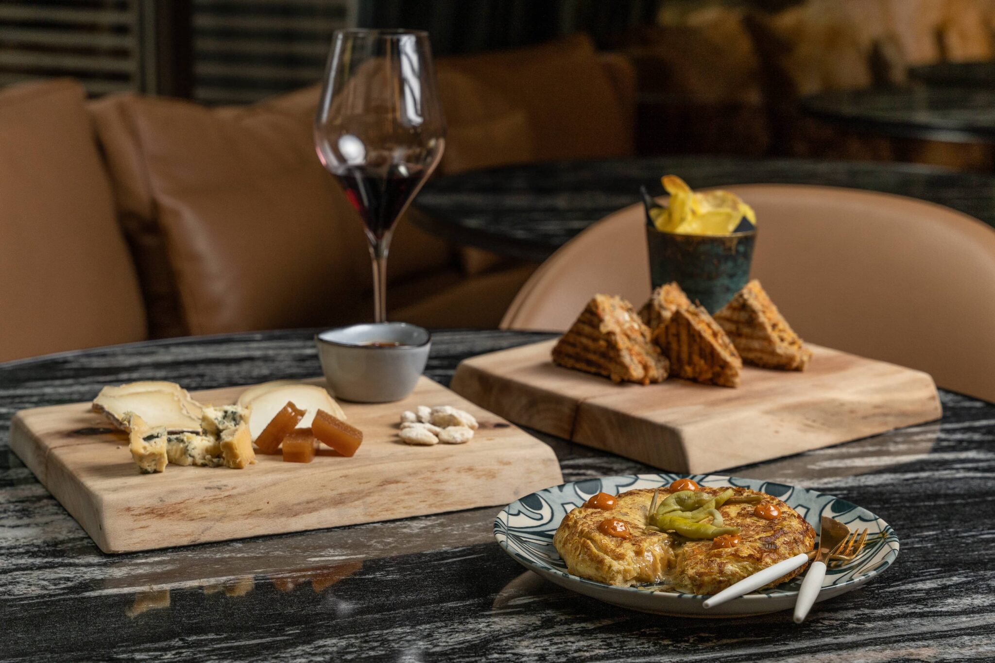 Una taula parada amb formatge, pasta de fruita, fruits secs, galetes, patates fregides, un plat de quiche i una copa de vi negre sobre taulers de fusta i un plat estampat.