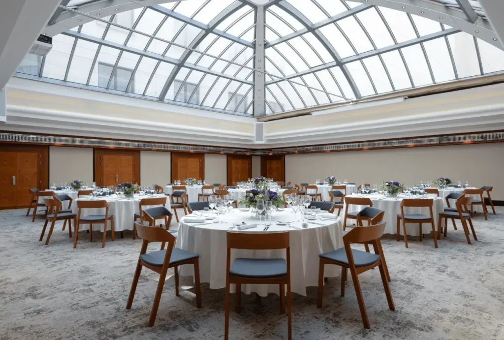 A spacious banquet hall at Alcron Prague with round tables set for an event, featuring a glass ceiling, wooden chairs, and floral centerpieces.