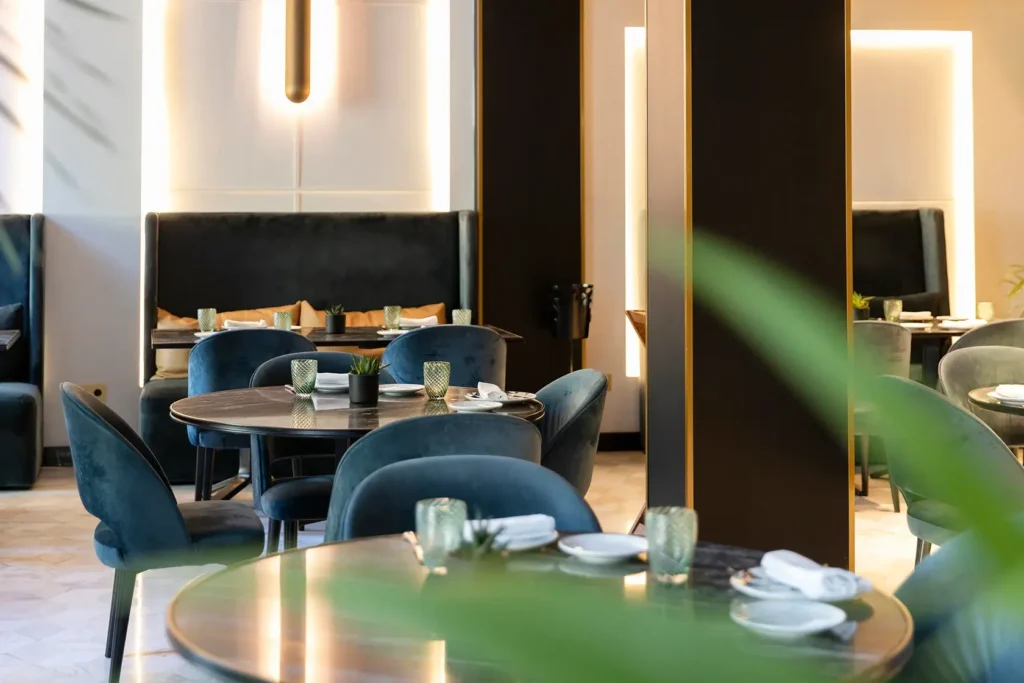 Modern Virens Restaurant interior with round tables, dark velvet chairs, neatly set tableware, and soft ambient lighting. Green leaves partially obscure the foreground.