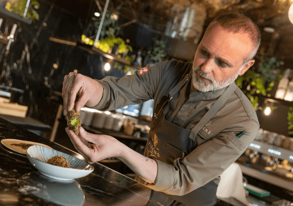 In the bustling kitchen of Virens Restaurant, a chef carefully plates food by hand, with a bowl and prepared ingredients in front of him.