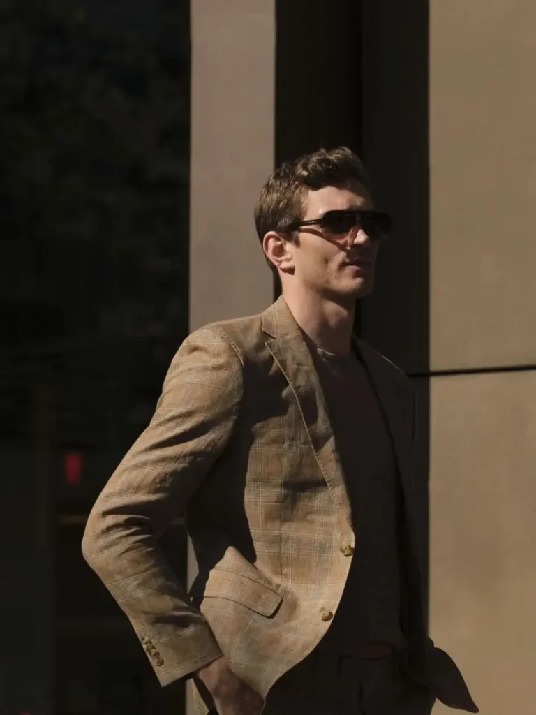 A man wearing sunglasses and a brown plaid suit stands outside near a building, enjoying the natural light as if on a stylish Shopping Escape, with his hands in his pockets.