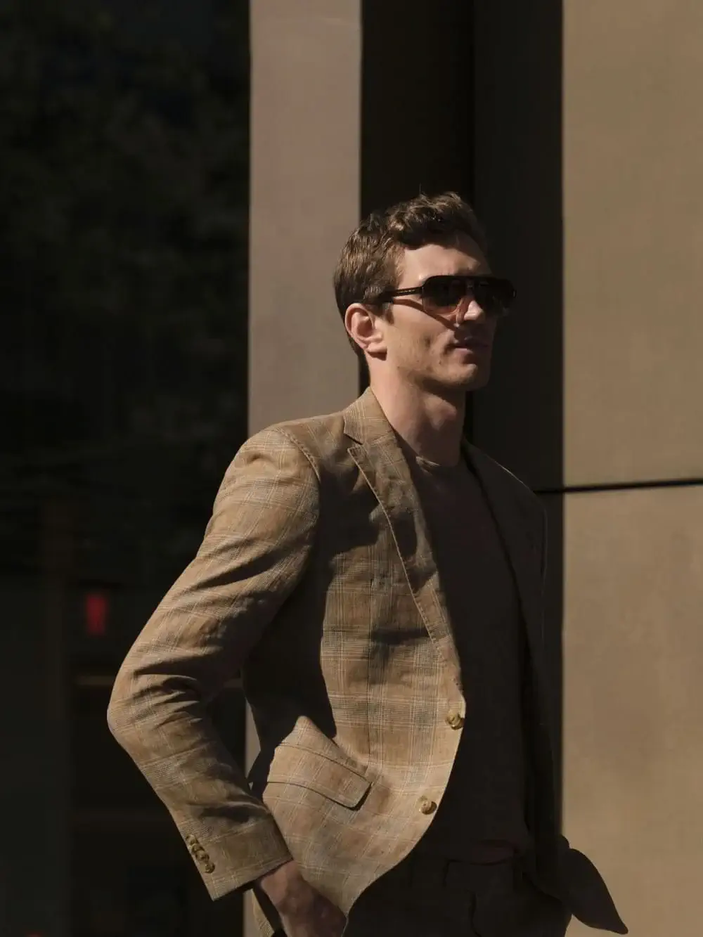 Un hombre con gafas de sol y un traje de cuadros escoceses se encuentra al aire libre con las manos en los bolsillos, parcialmente iluminado por la luz del sol, listo para una escapada de compras.