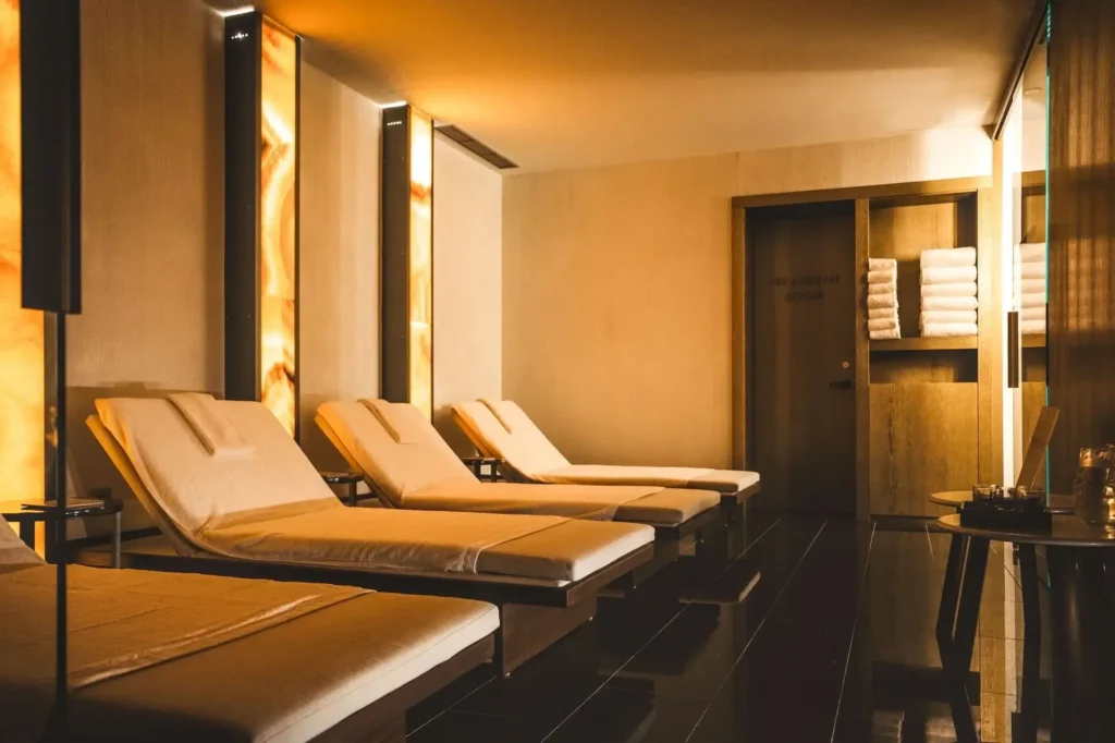 An Inspired Spaces dimly lit spa relaxation room with four cushioned lounge chairs, glowing wall panels, stacked towels on shelves, and a door labeled "Hydro Pool & Sauna.