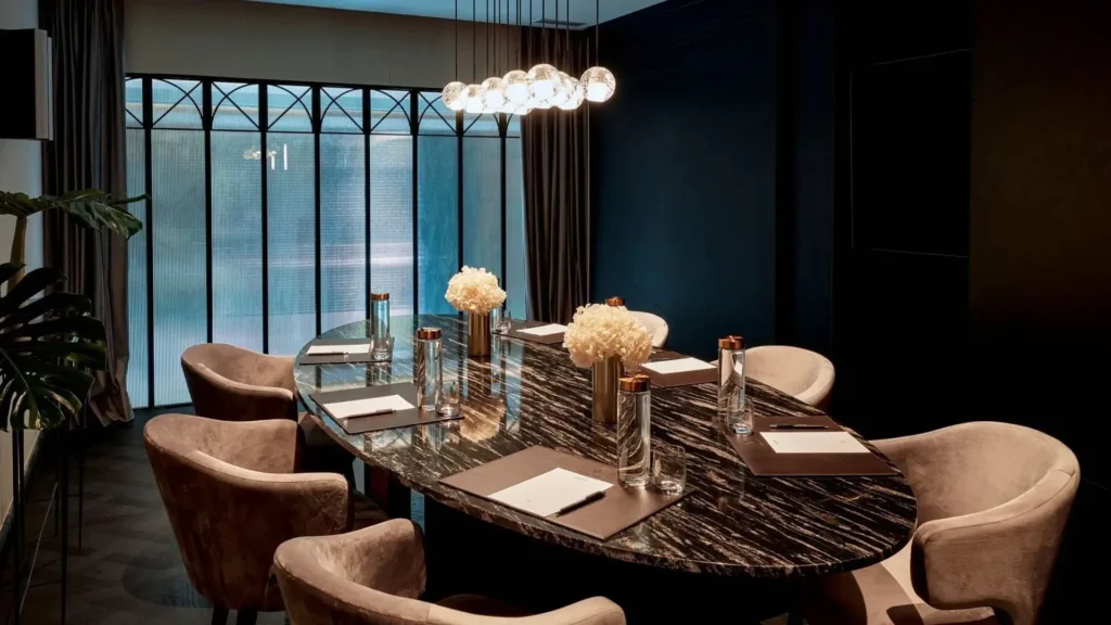 Una moderna sala de conferencias con una mesa de mármol negro, sillas beige, blocs de notas, bolígrafos, jarrones de flores y botellas de agua, iluminada por lámparas colgantes cerca de grandes ventanales.