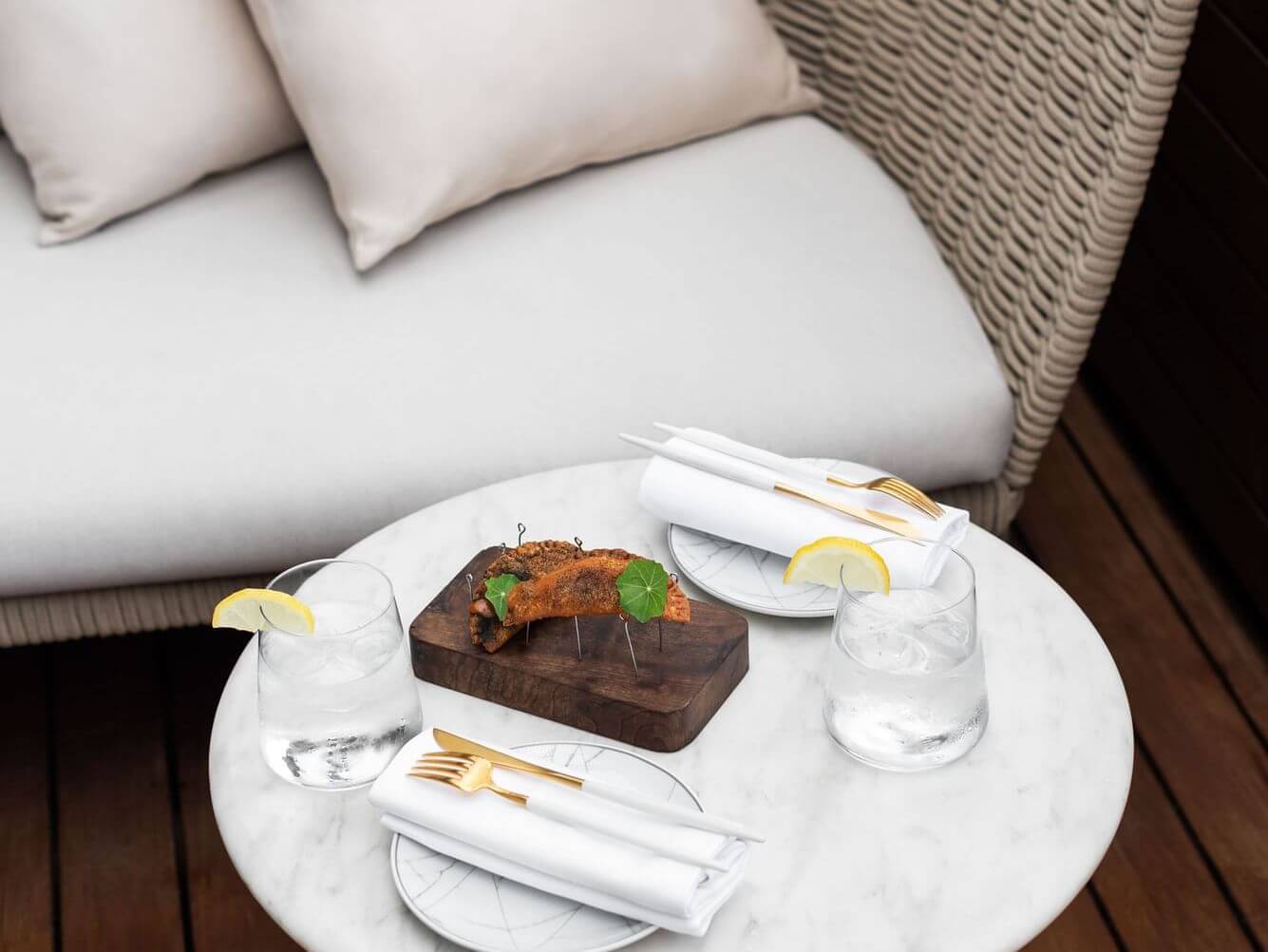 Una taula de marbre a la suite Penthouse té dos gots d'aigua amb rodanxes de llimona, plats amb coberts embolicats amb tovallons i una petita tauleta de fusta amb menjar, tot col·locat davant d'un sofà de vímet encoixinat.