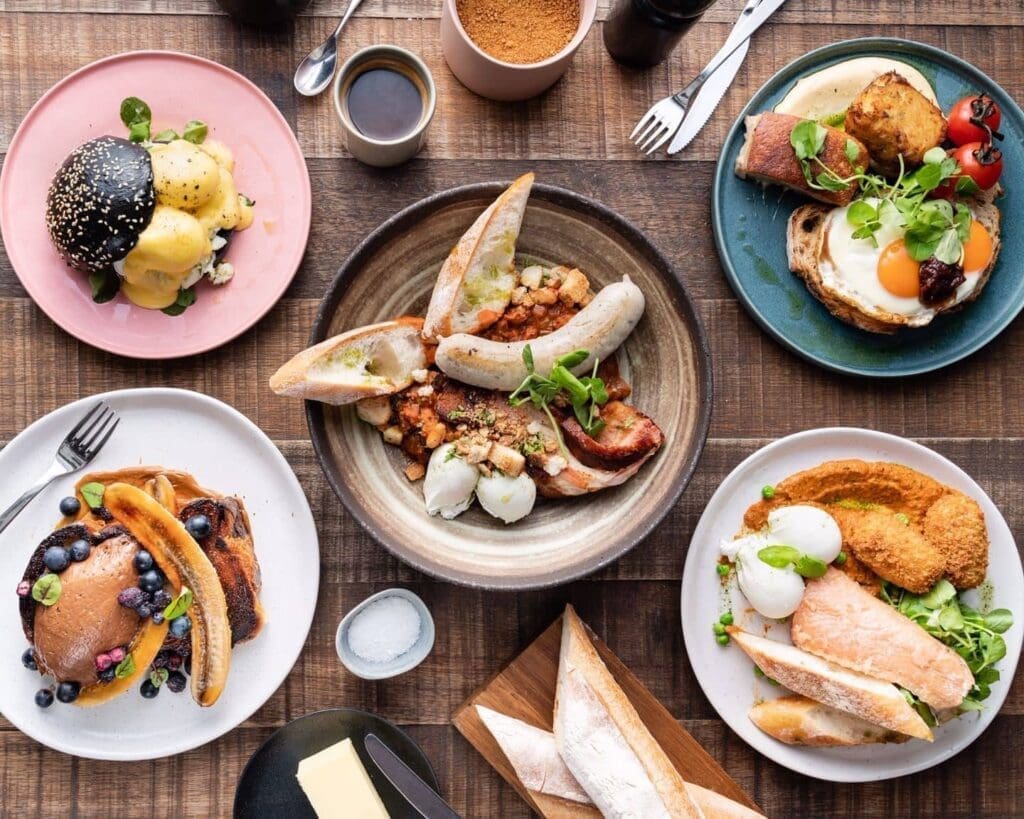 A table set with assorted breakfast dishes, including eggs, sausages, toast, and pancakes with blueberries at Hardware Société Barcelona, alongside grilled vegetables and coffee on a wooden surface.