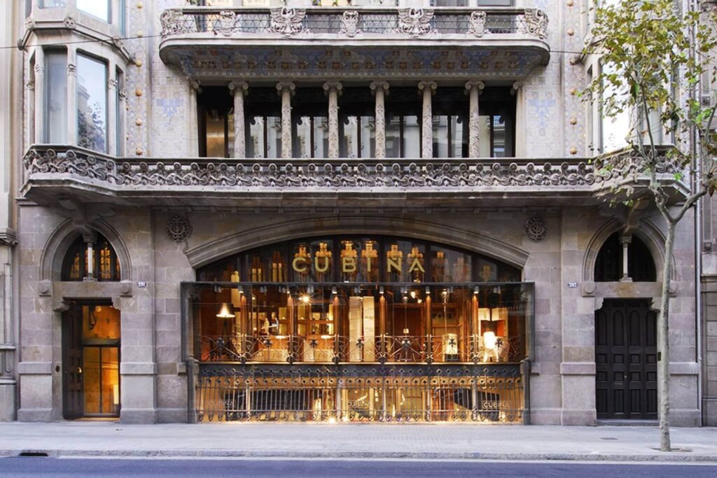 Un edificio de piedra de Barcelona con detalles arquitectónicos ornamentados y grandes ventanas arqueadas luce el letrero dorado "CUBIÑA" sobre su entrada; la cálida iluminación interior se deja ver a través del cristal, evocando la elegancia del cercano castillo de Montjuïc.