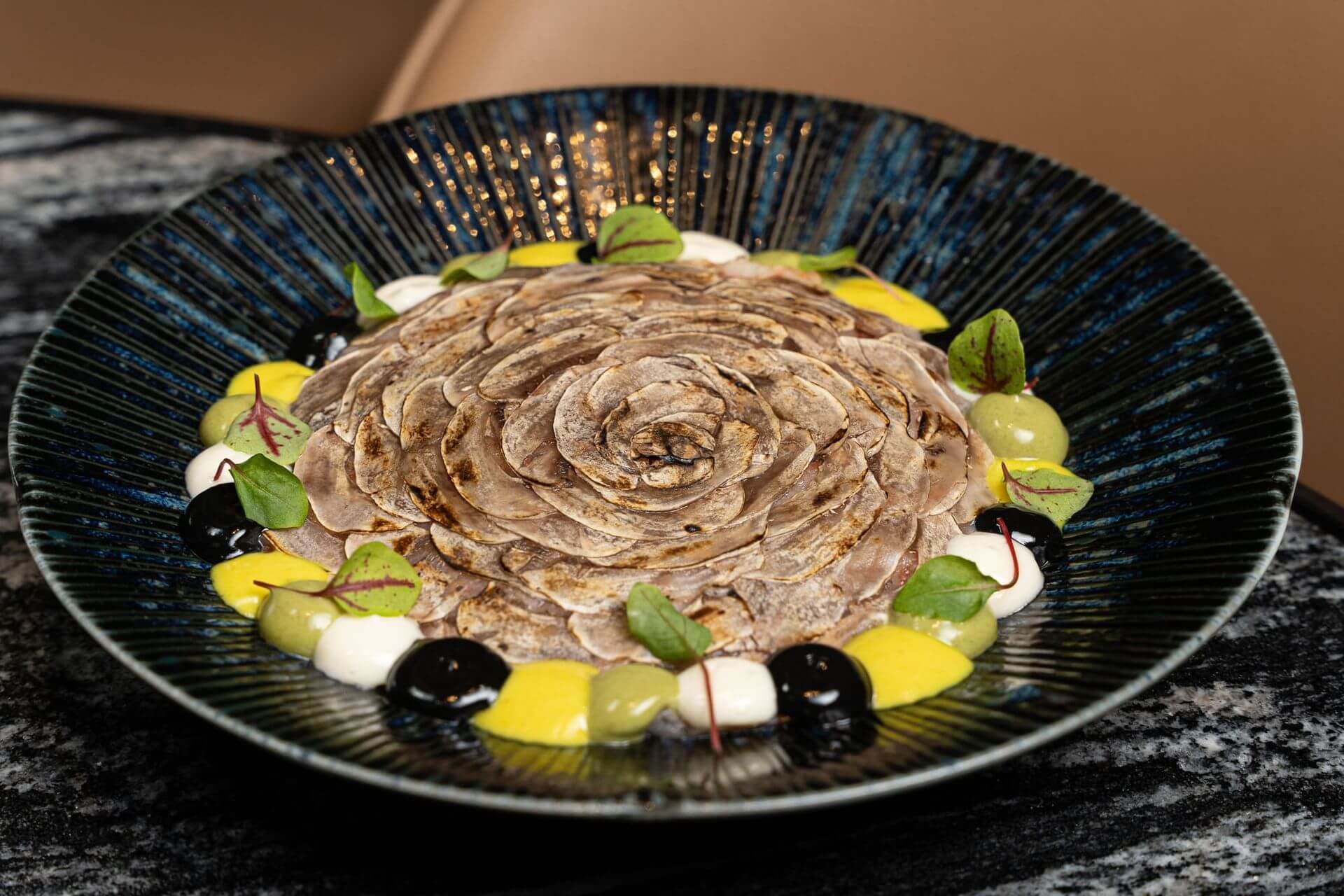 A dark bowl with thinly sliced mushrooms arranged in a flower pattern, surrounded by colorful dollops of sauces and garnished with small green leaves.